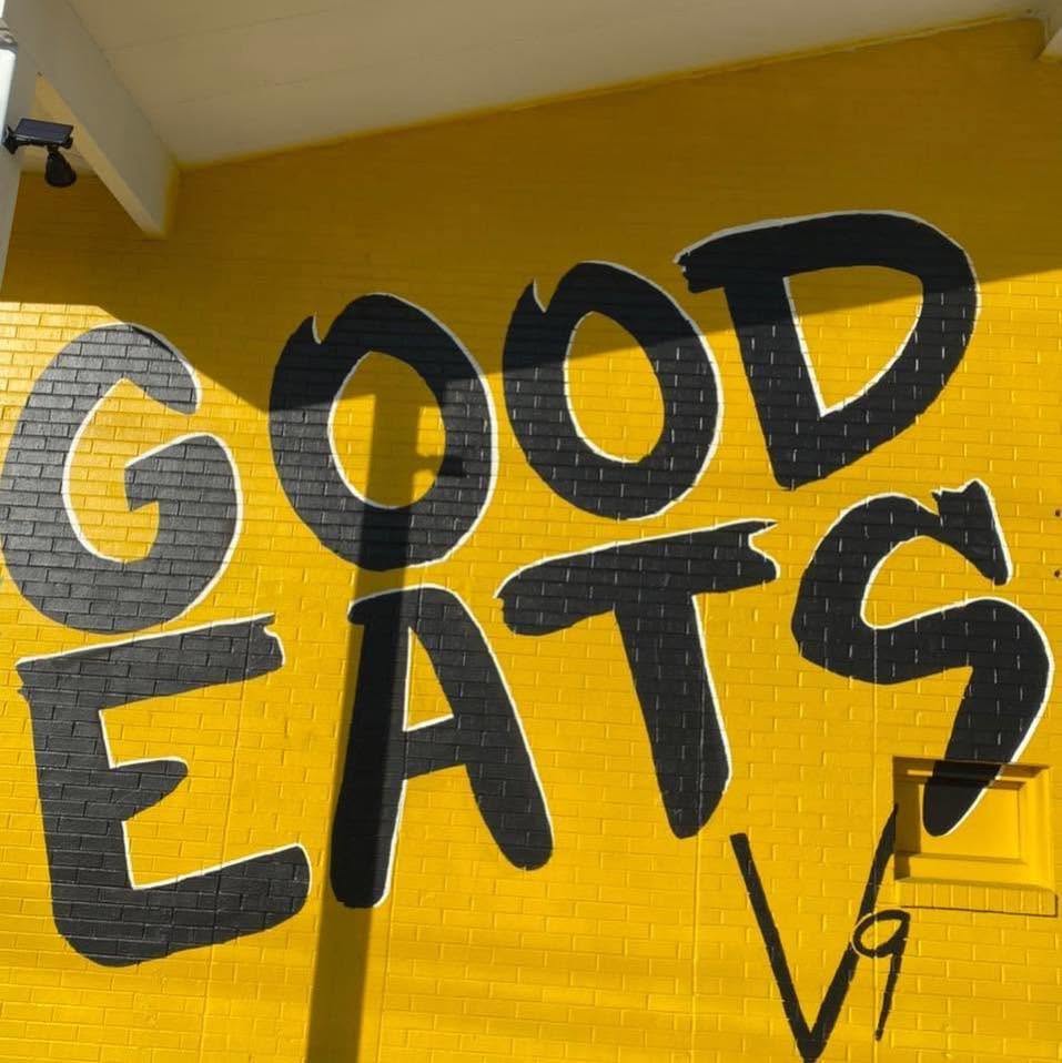 The hand-painted GOOD EATS mural in black on the sunflower-yellow wall, signed V9