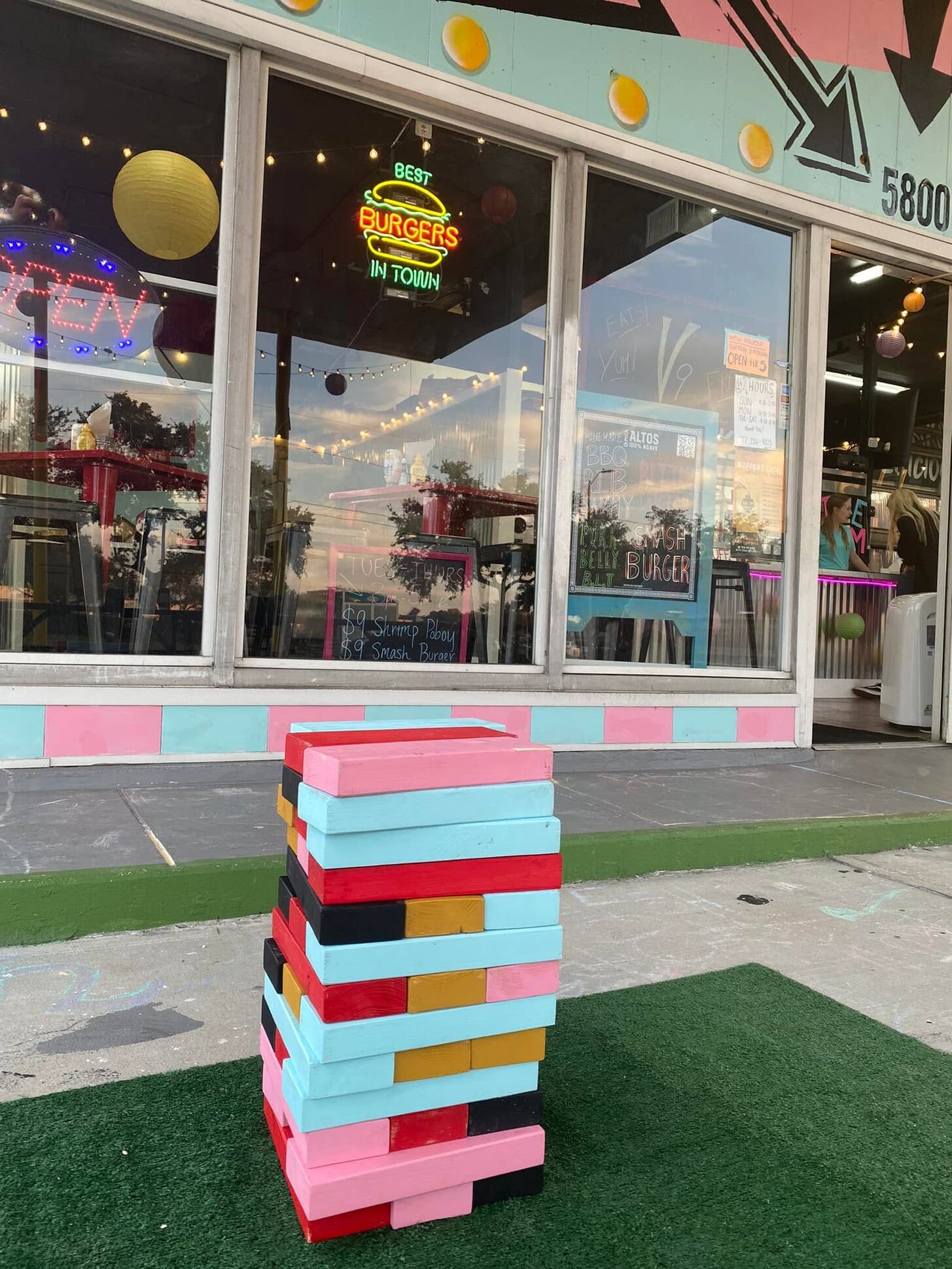 The garage-bay windows of V9 EATS with the BEST BURGERS IN TOWN neon sign and a giant painted block-stacking game on the turf